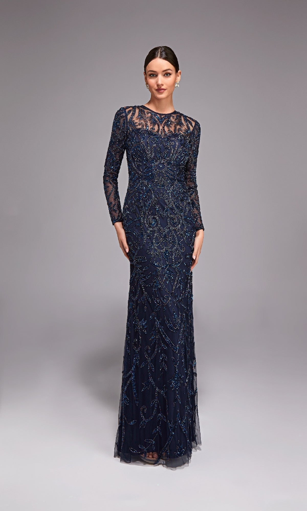 A woman poses gracefully in front of a plain, light gray background wearing the Alyce 27778 long formal dress in Midnight—a beaded evening gown with long sleeves and a sheer neckline.